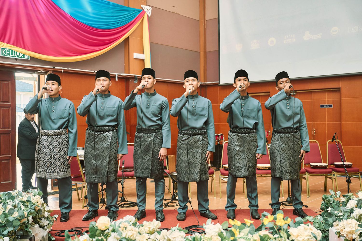 Six Malaysian men in matching teal Baju Melayu saluting during a formal indoor cultural performance.