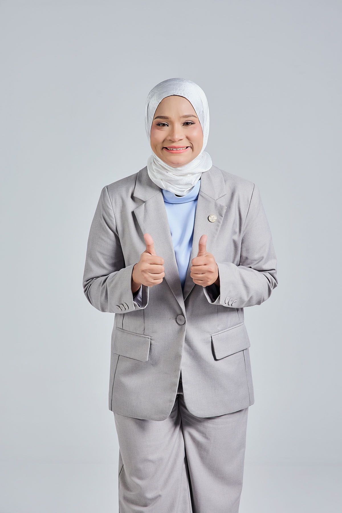 Smiling Muslim businesswoman in gray suit and white hijab giving thumbs up gesture on gray background.