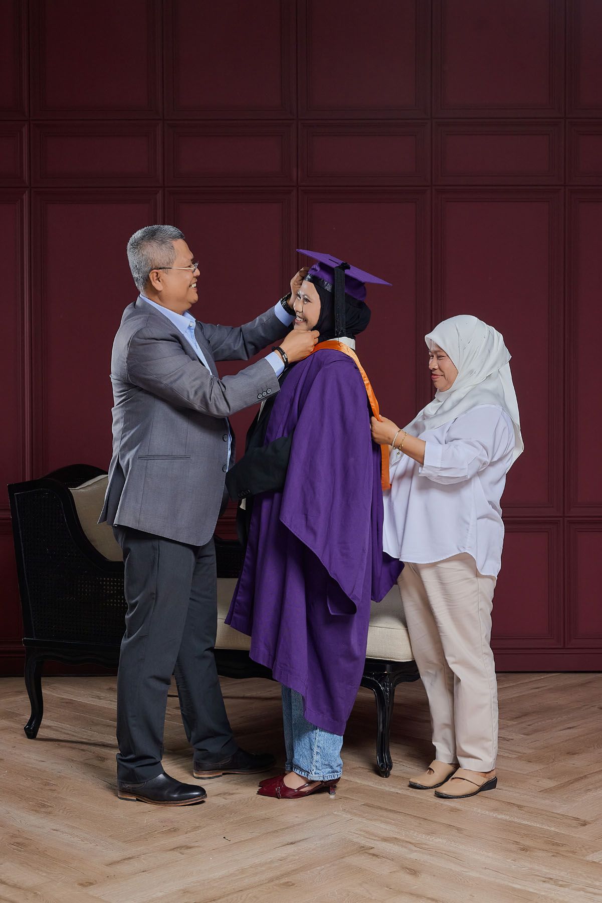 Parents proudly adjusting graduation cap and gown on their daughter in elegant red room.