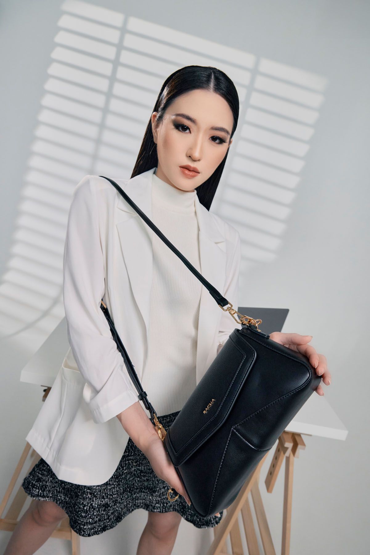 Asian woman in white blazer and tweed skirt holding a black structured crossbody bag.