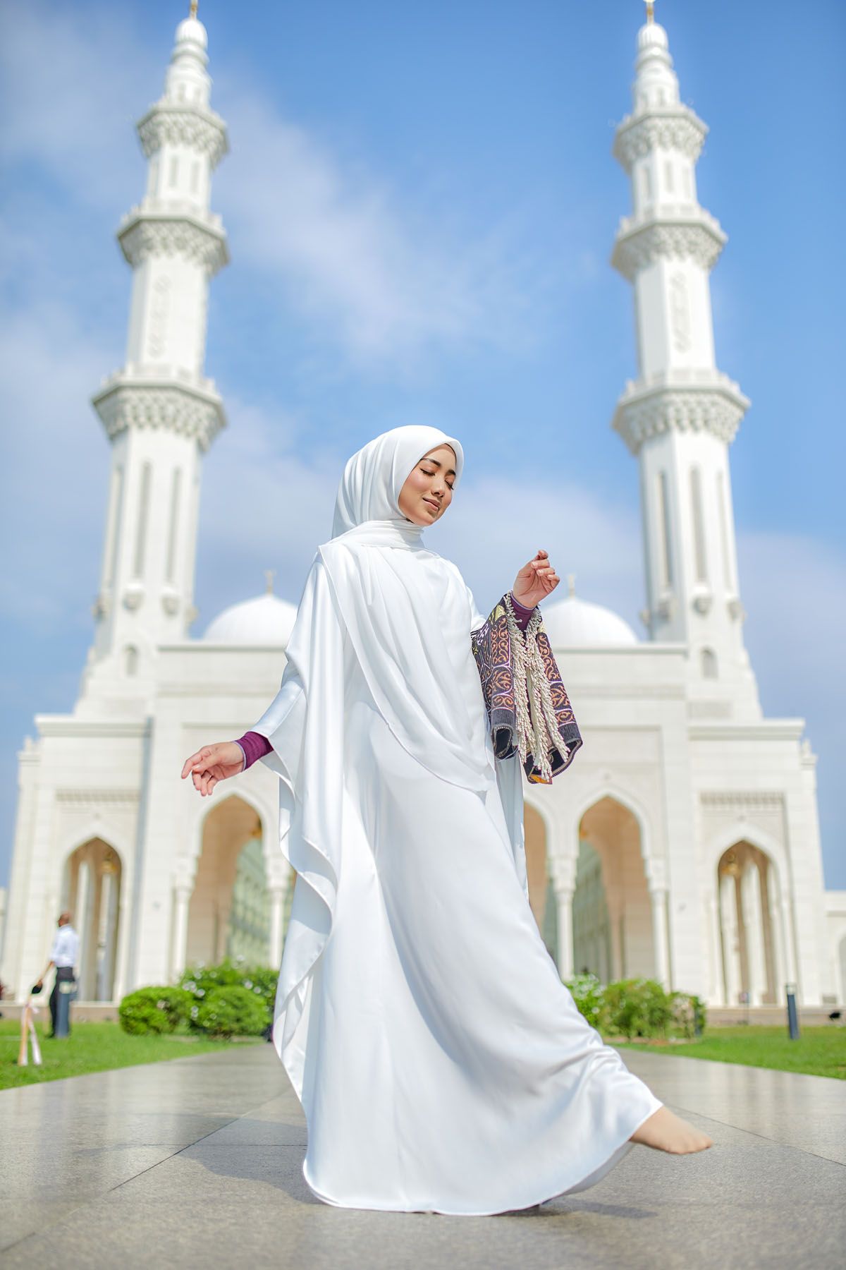 Muslim woman in white hijab and abaya walking gracefully in front of a stunning white mosque with tall minarets.