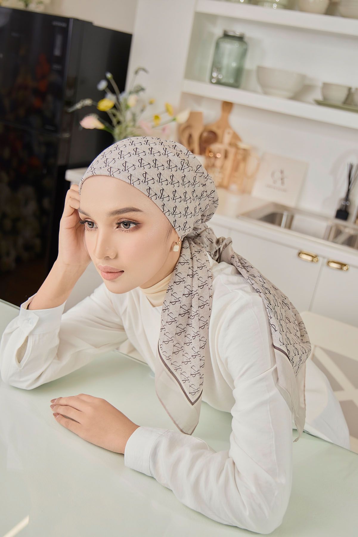 Woman in white blouse wearing beige printed hijab poses thoughtfully in modern kitchen.