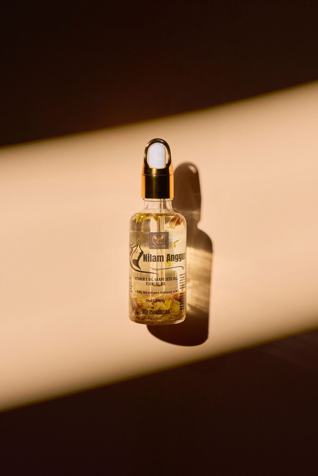 Gold dropper serum bottle by Nilam Anggur floating against a warm brown background with dramatic shadows.