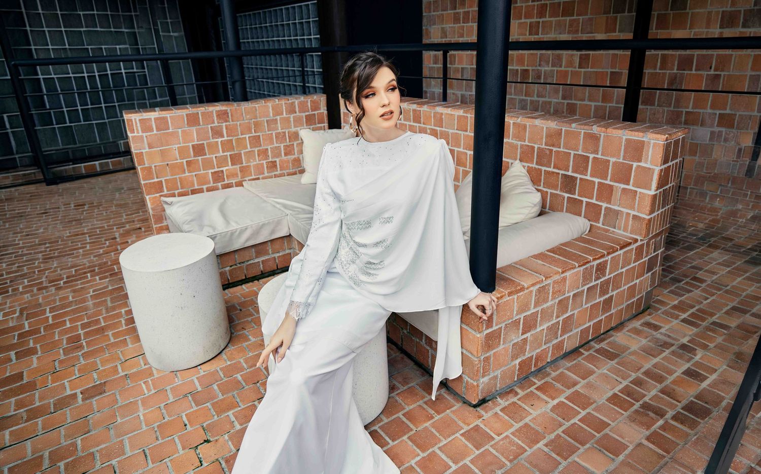 Woman in white modest long-sleeve dress and cape sits elegantly on brick outdoor bench.