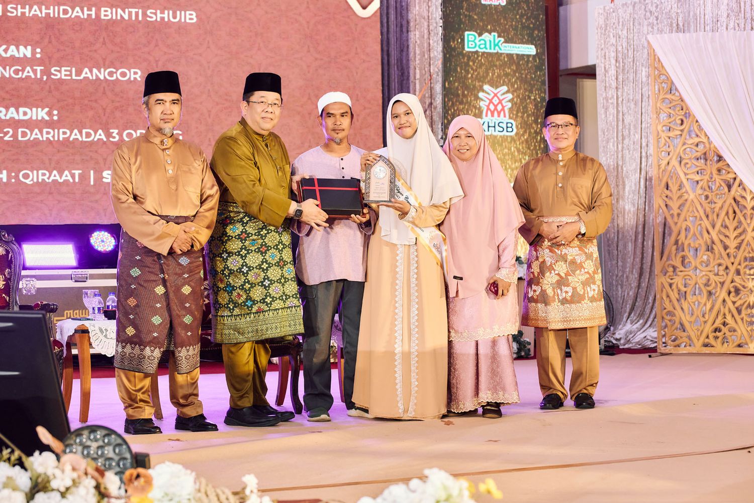 Muslim woman receives award on stage at a formal Quran recitation ceremony in Malaysia with officials in traditional attire.