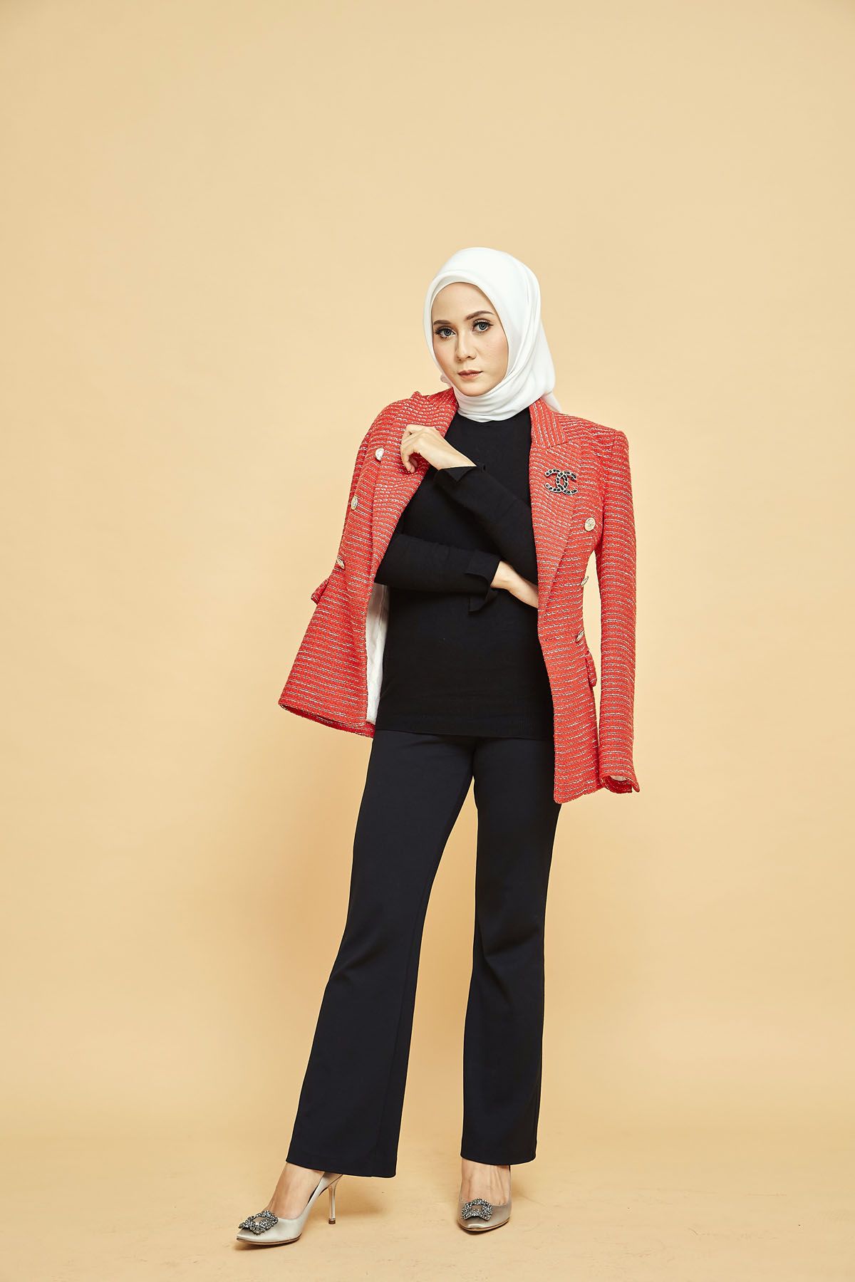 Muslim woman in white hijab adjusting red tweed blazer while posing on a beige background.