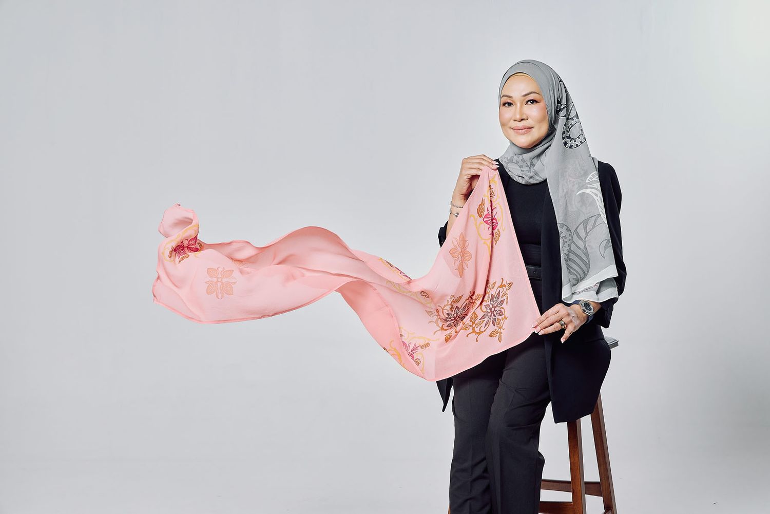 Muslim woman in a gray hijab and black outfit holding a flowing pink floral scarf on a stool.