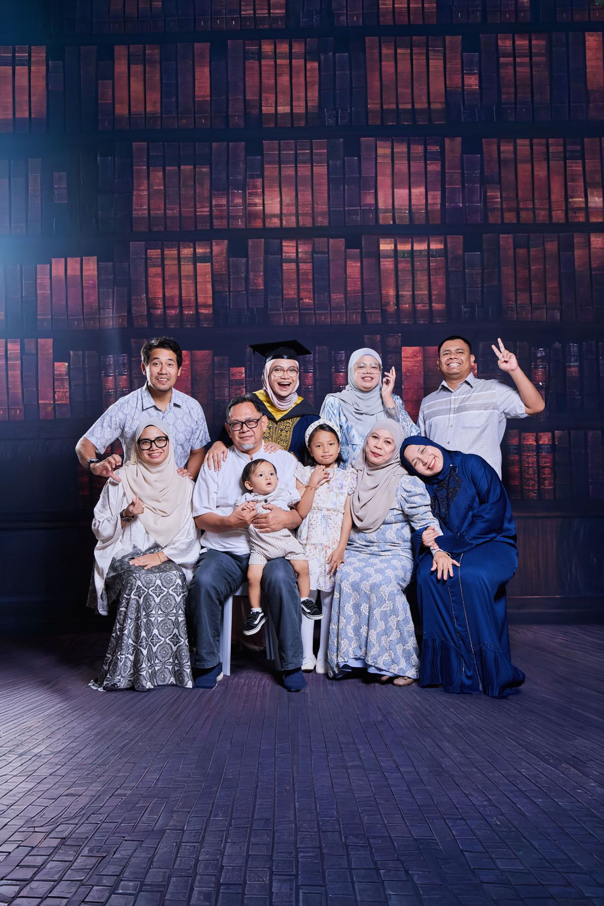 Large family graduation portrait with graduate in cap and gown surrounded by nine relatives before bookshelf backdrop.