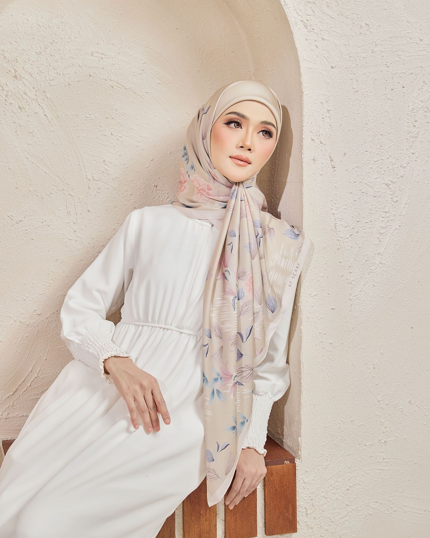 Woman in white dress wearing beige floral printed hijab scarf leaning against archway wall.