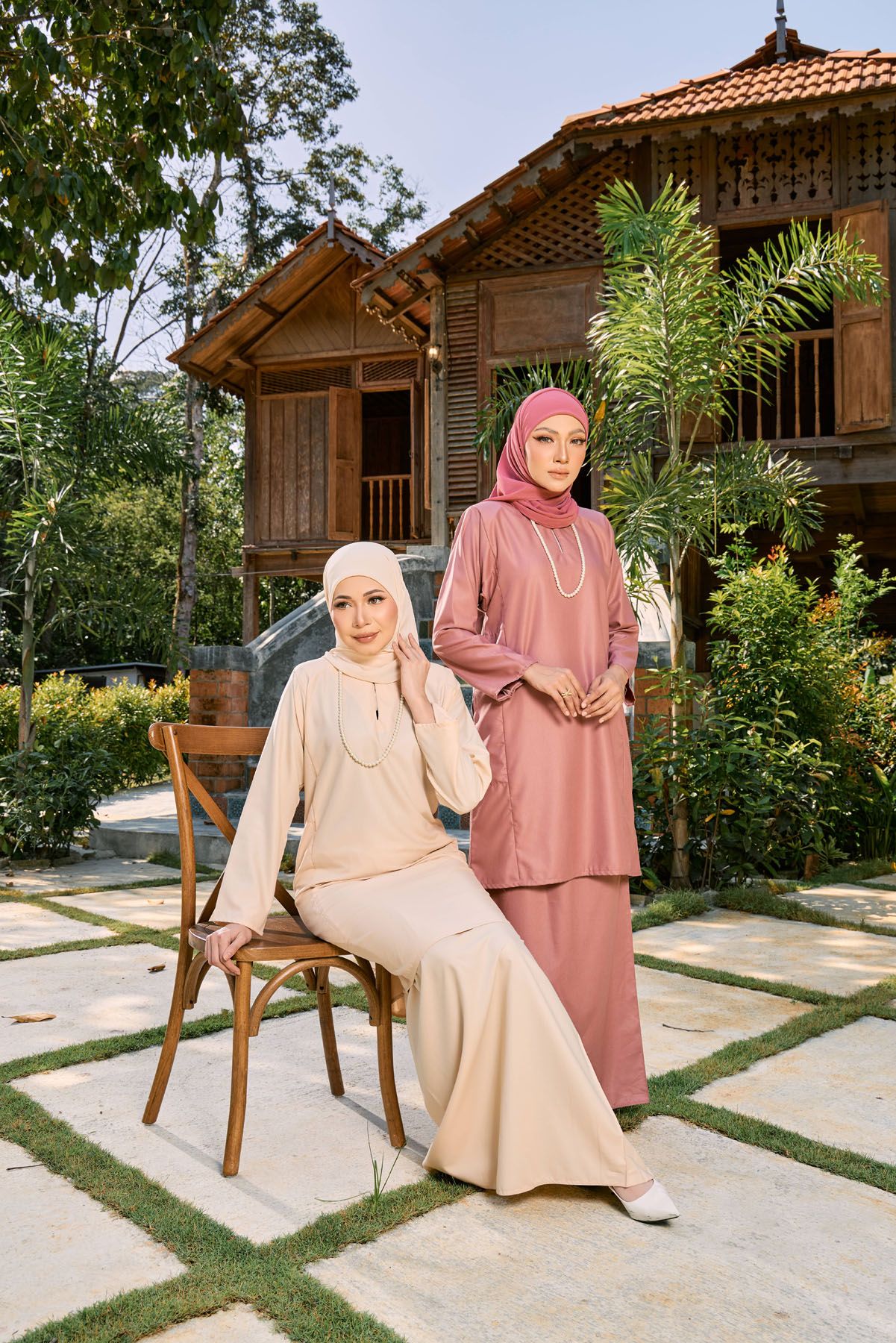 Two women in dusty pink and beige baju kurung with pearl accessories posing outside traditional house.