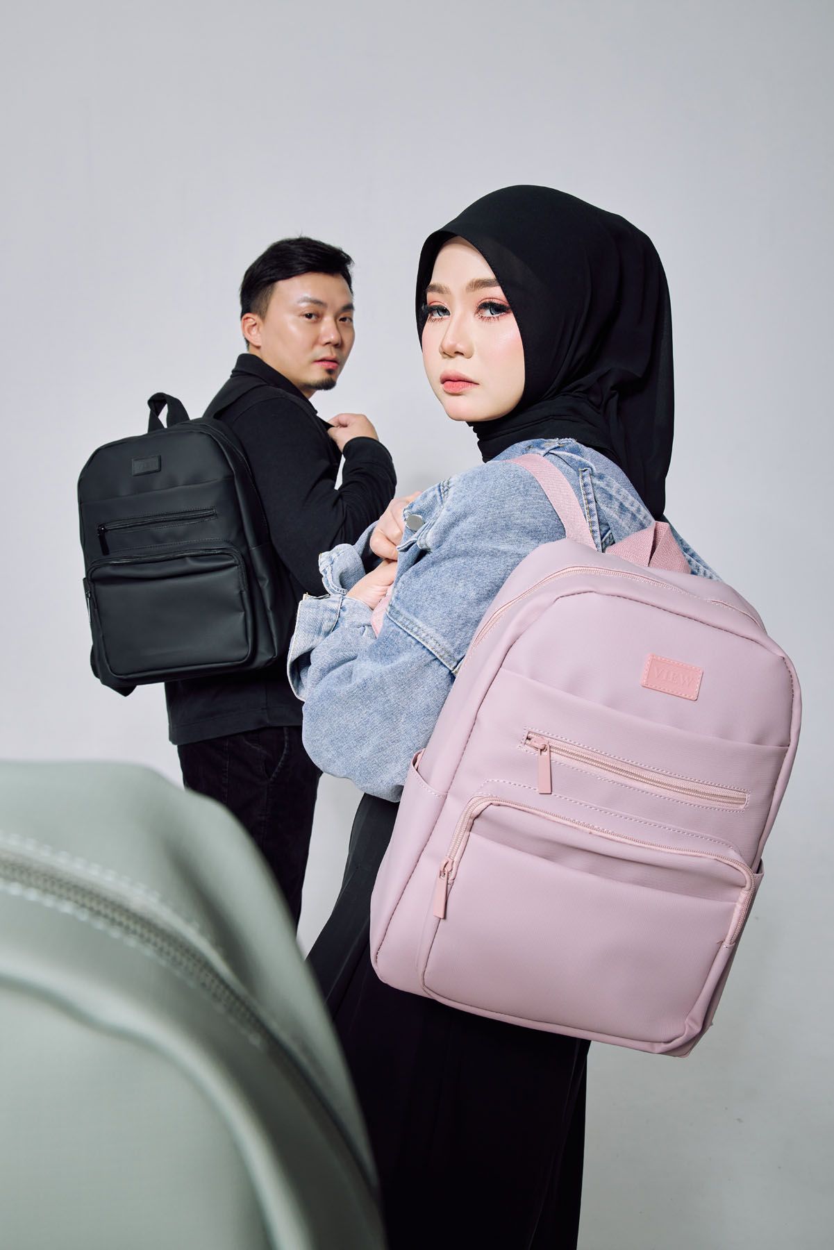 Man and woman in hijab modeling black and pink backpacks against a light gray studio background.