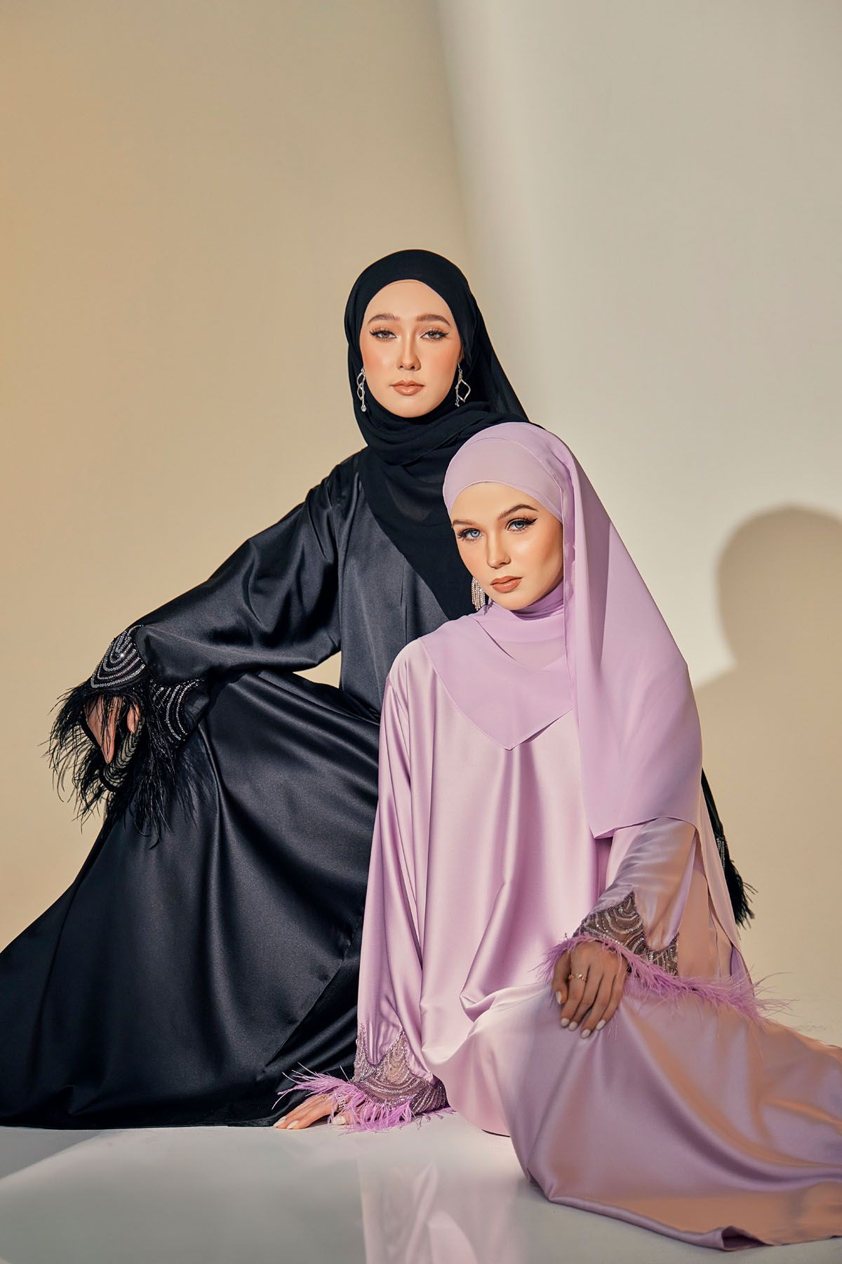 Two women in black and lilac feather-trimmed satin hijab abayas posing on a beige studio background.