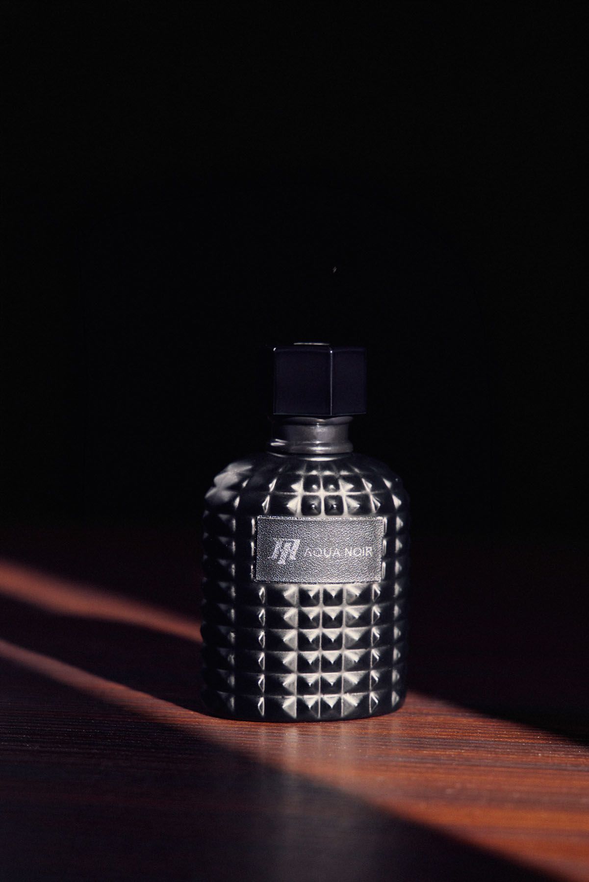 Dark studded Aqua Noir fragrance bottle with pyramid texture on wooden surface against black background.