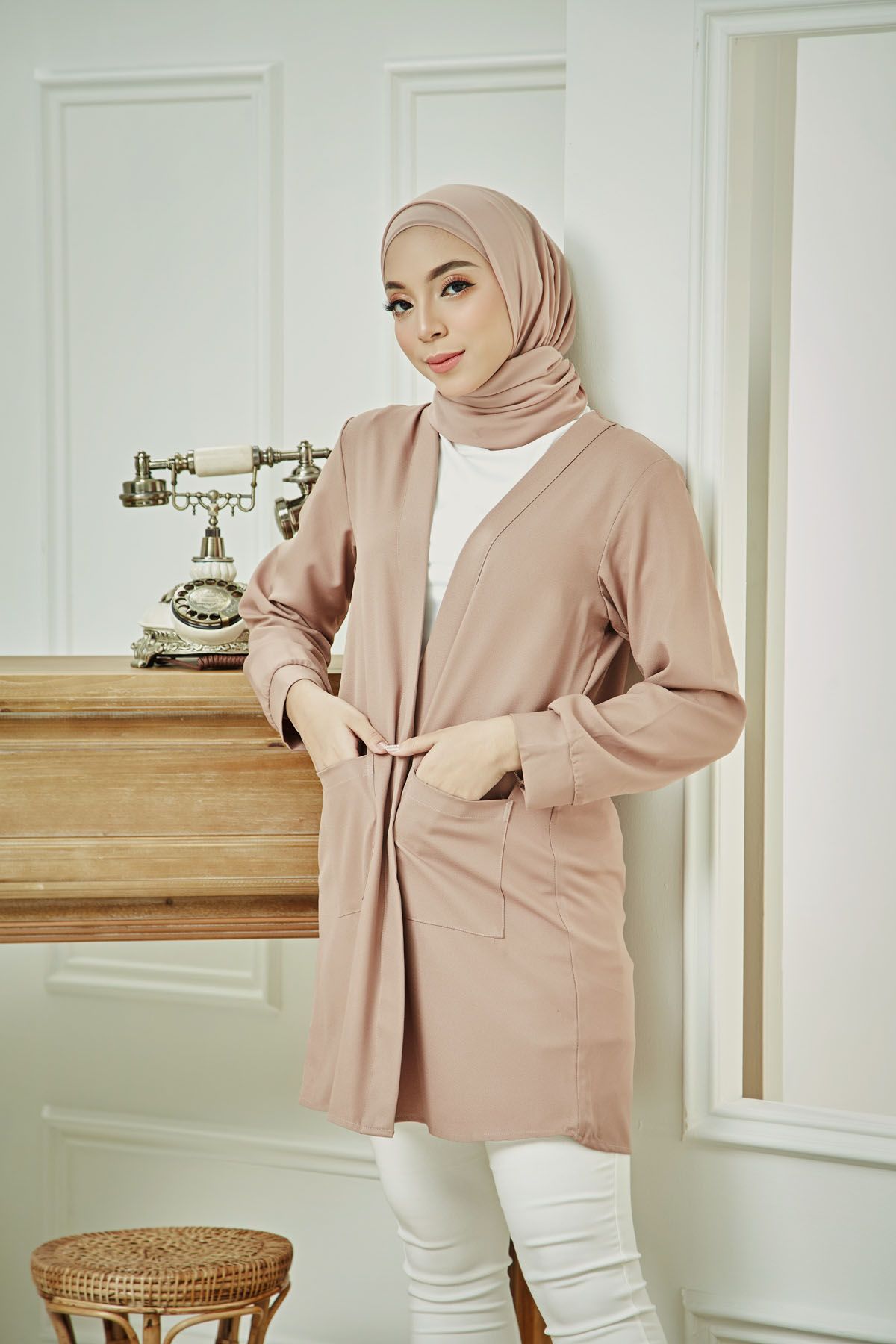 Woman in dusty pink cardigan and matching hijab posing near vintage telephone on wooden shelf.