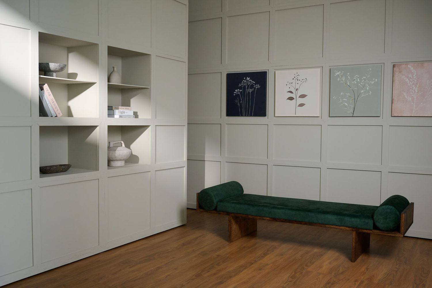 Modern minimalist room with white built-in shelving, green velvet bench, hardwood floors, and botanical art prints on wall.