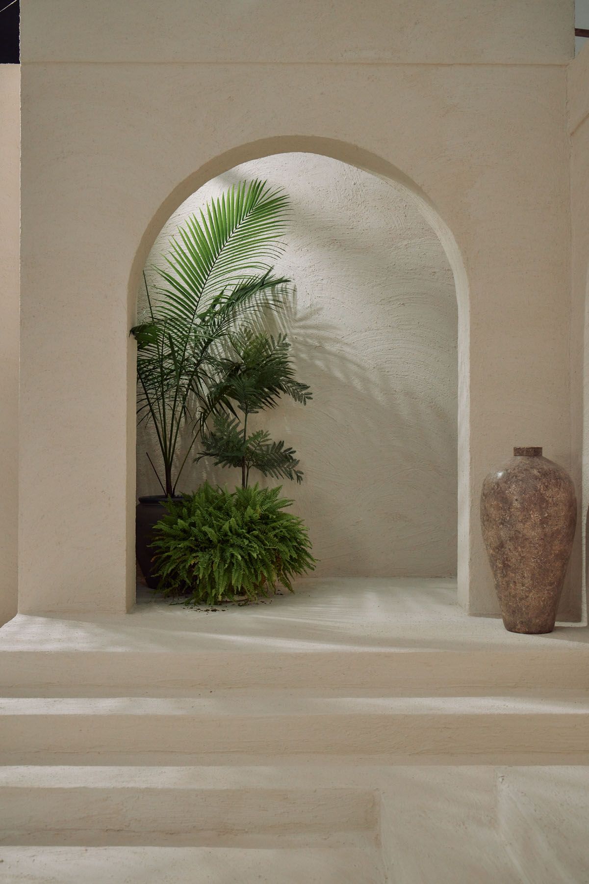 White plaster arched niche with tropical palm and fern plants beside a rustic clay vase.