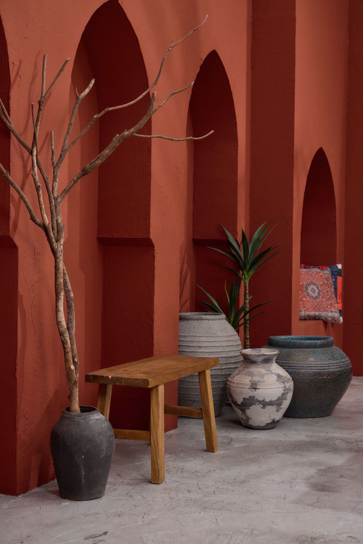 Rustic terracotta courtyard with arched niches, bare branch tree, wooden stool, and large ceramic pottery vessels.