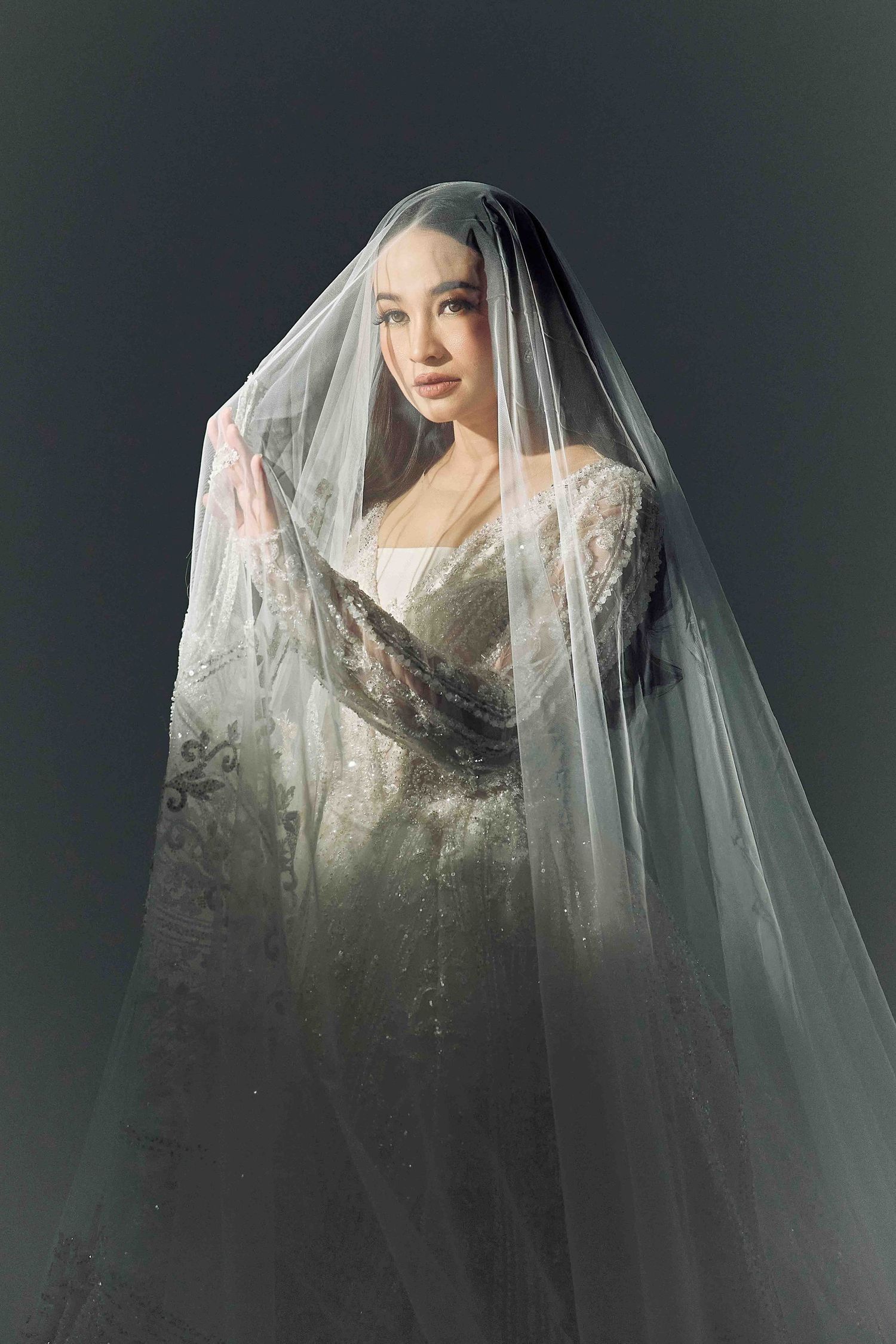 Bride in embellished lace gown lifts a sheer veil dramatically against a dark studio backdrop.