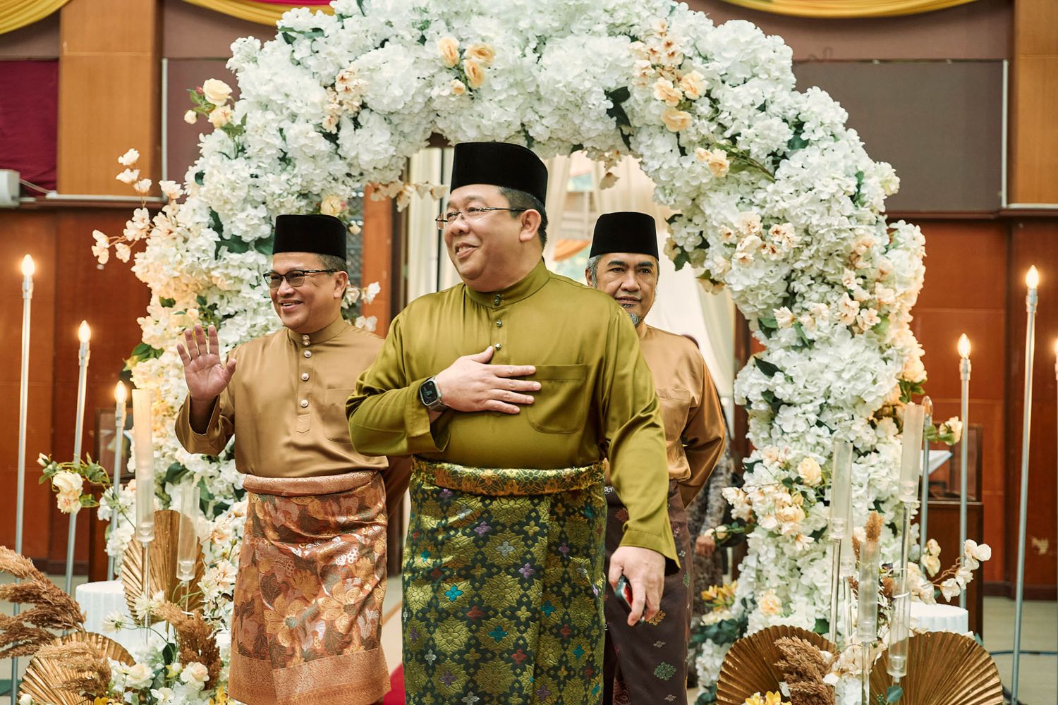 Smiling Malaysian man in olive Baju Melayu waving under a white floral arch at a formal celebration.