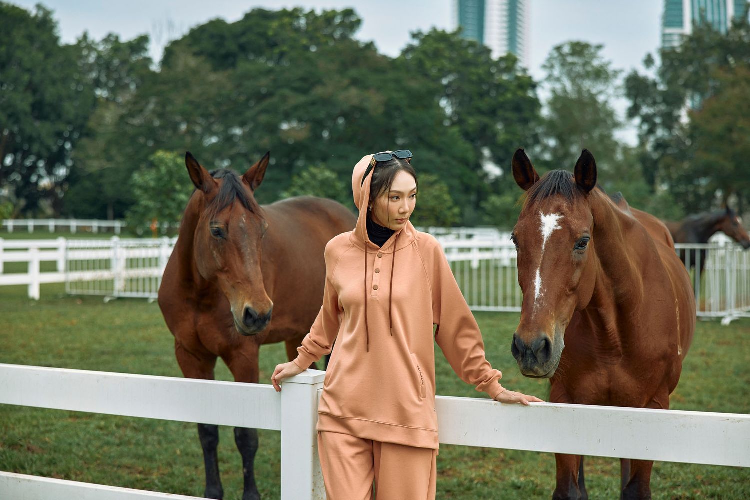 Woman in peach hijab outfit standing beside two brown horses at white fence paddock.