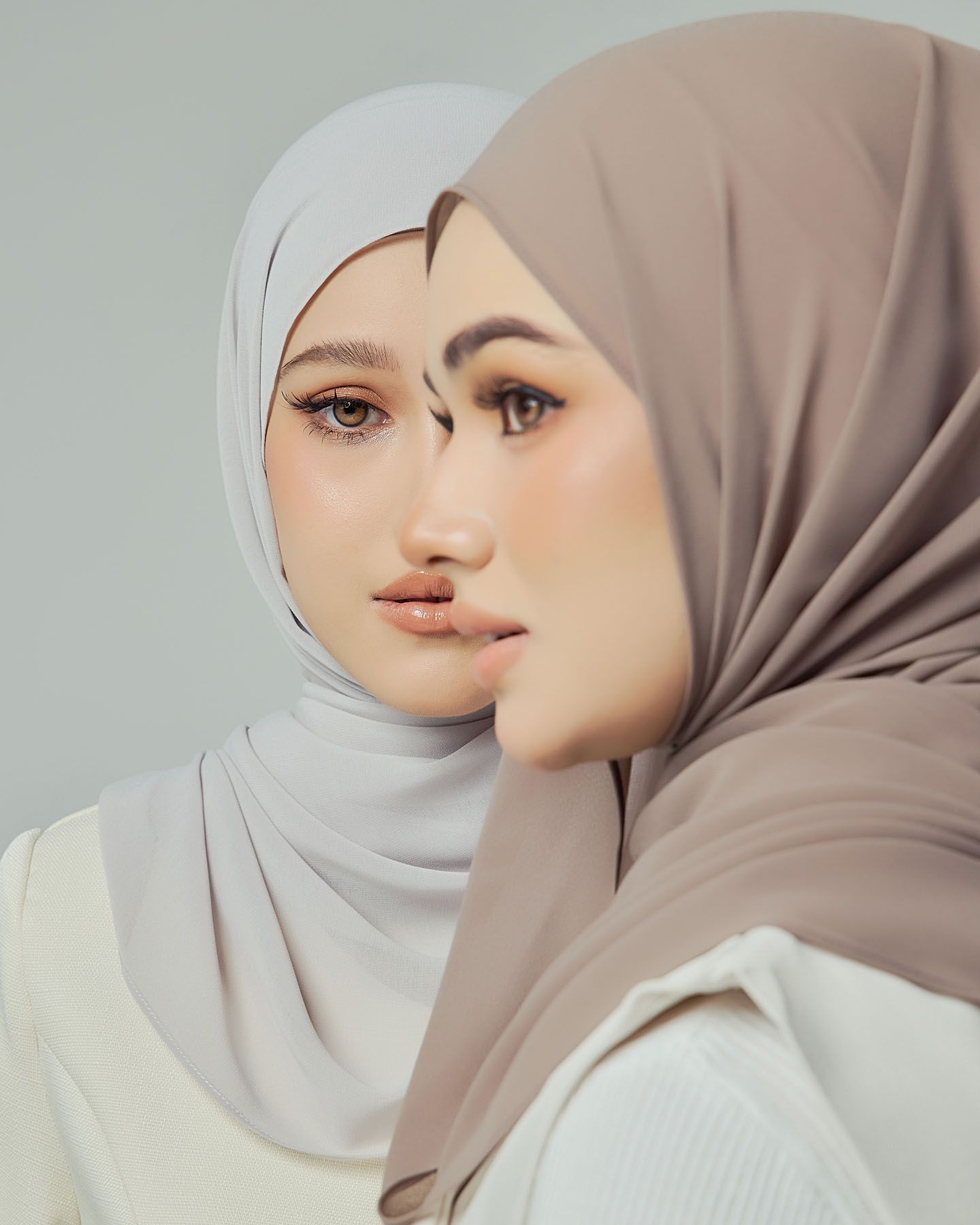 Two women in taupe and grey chiffon hijabs with white outfits in elegant close-up studio portrait.