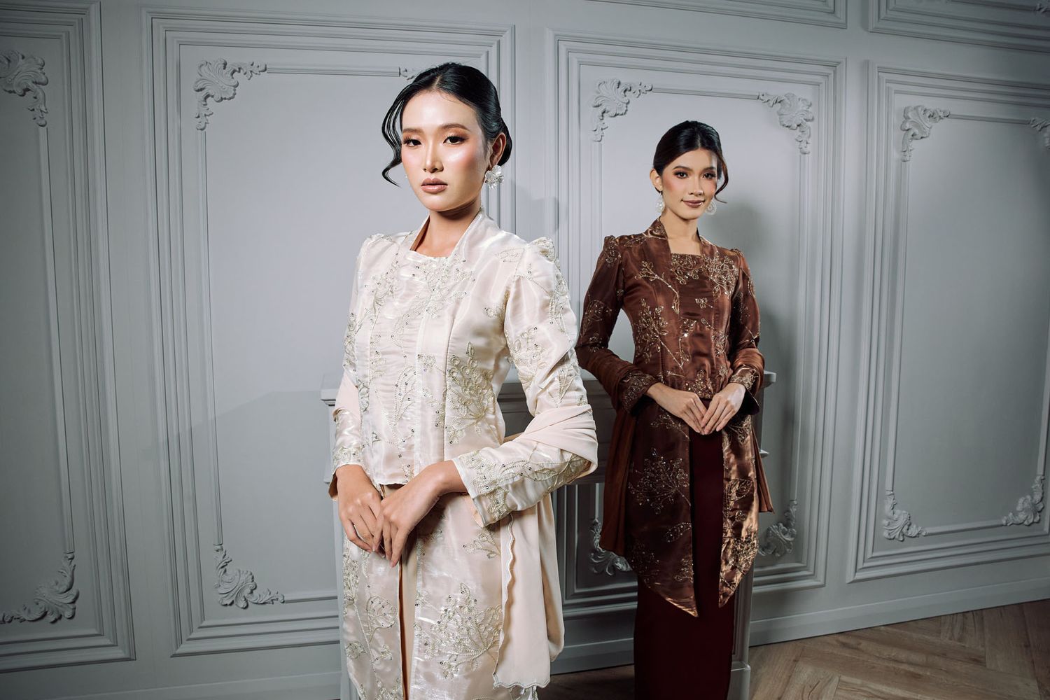 Two women modeling traditional kebaya outfits, one in white lace and one in brown velvet, in an elegant interior.