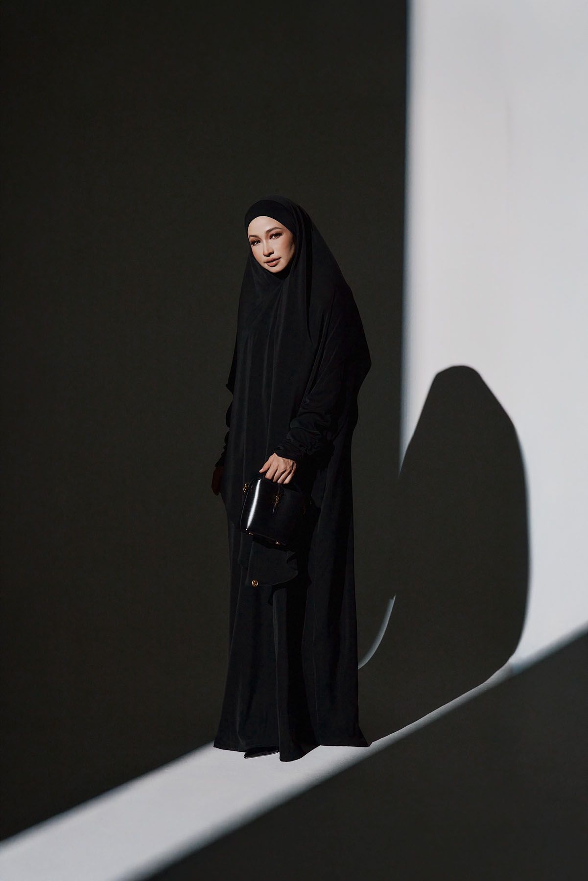 Woman in all-black abaya and hijab stands holding a book against a dramatic light and shadow background.