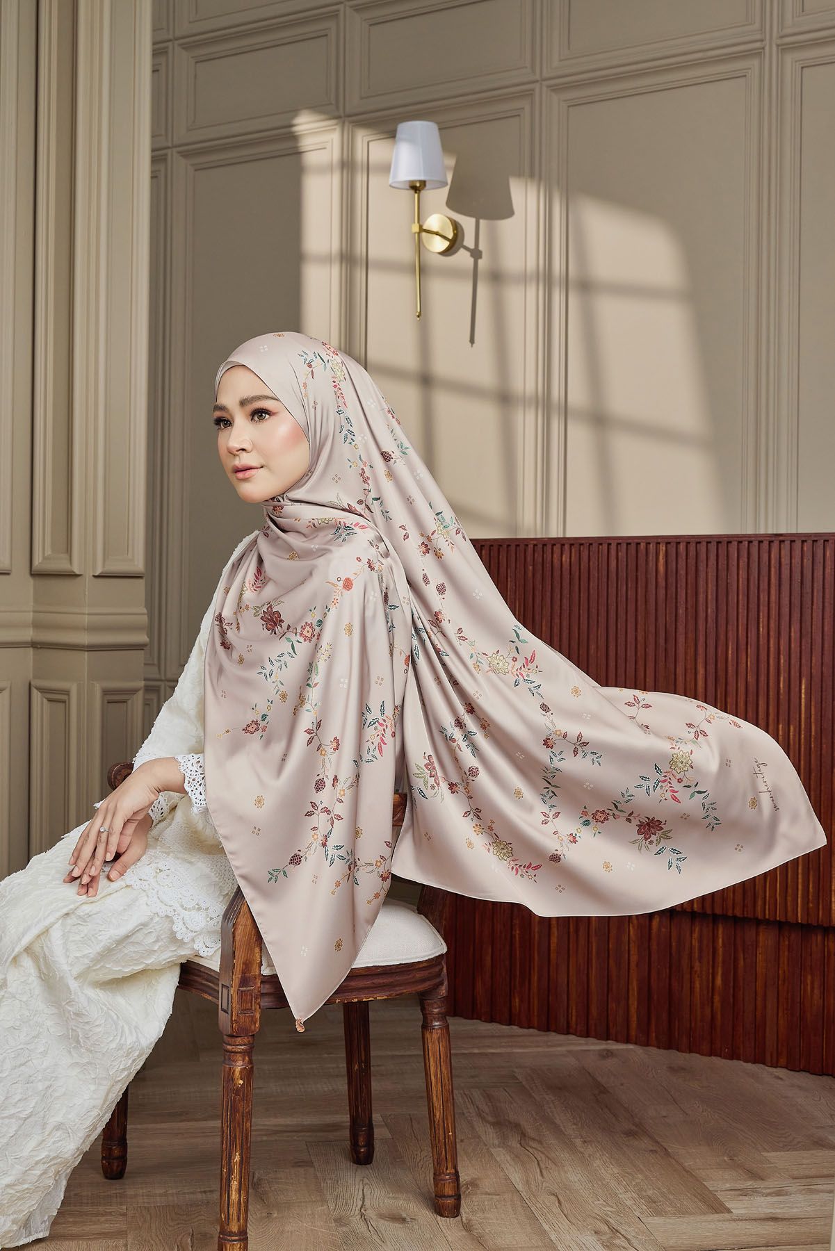 Woman in floral pink hijab and white lace baju kurung seated gracefully on a wooden chair.