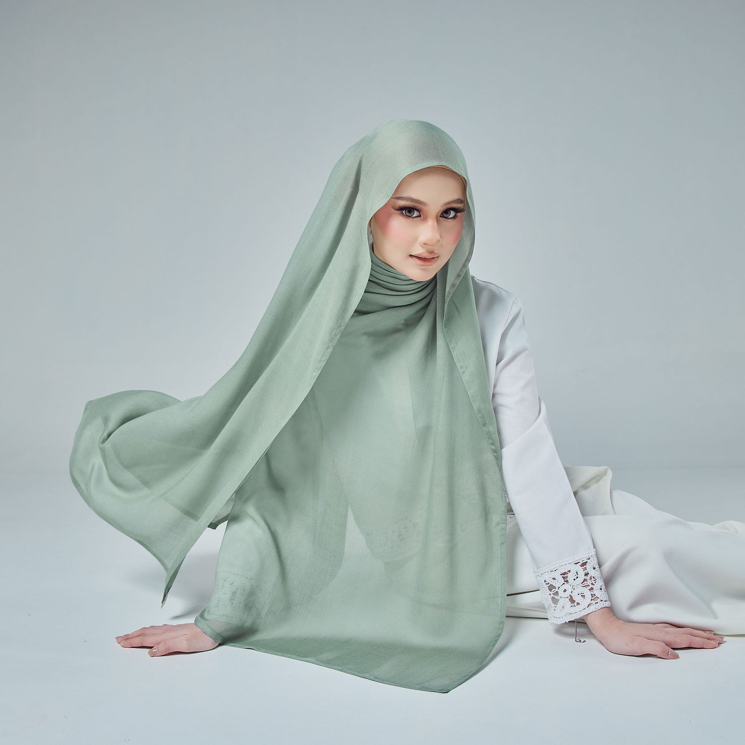 Woman in mint green hijab and white lace dress sits gracefully on light studio floor.