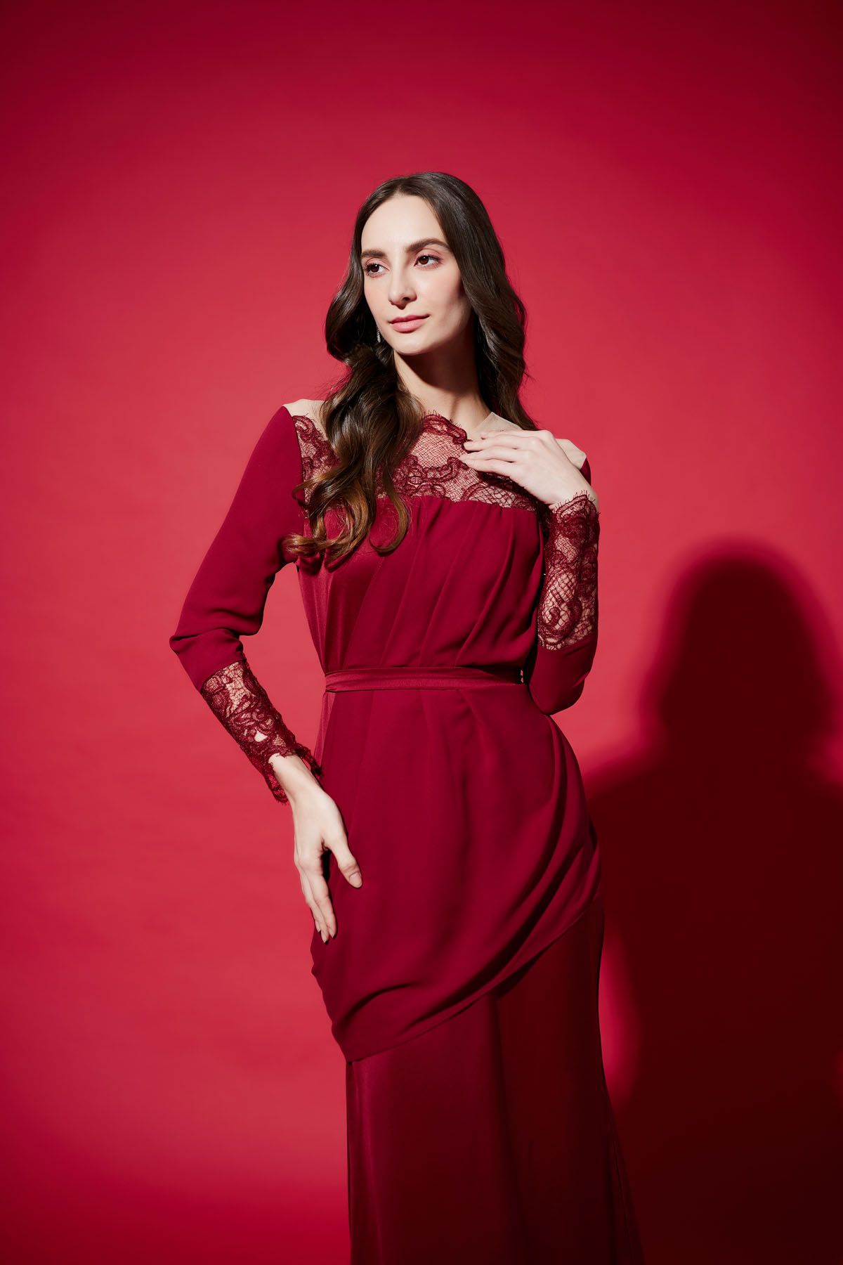 Sophisticated woman in a deep red lace-detail evening gown posing against a bold red background.