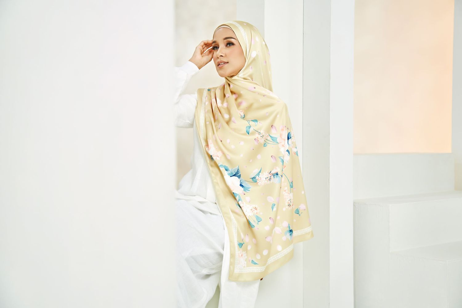 Stylish Muslim woman in cream floral hijab leaning against white wall in bright minimalist interior.