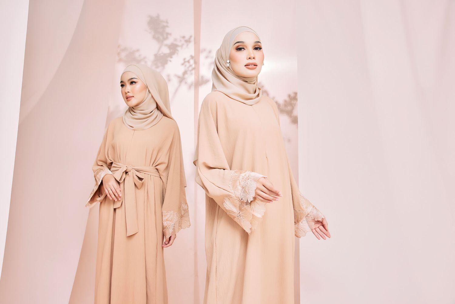 Two women in matching beige hijab and abaya with lace cuffs pose in a soft pink studio.