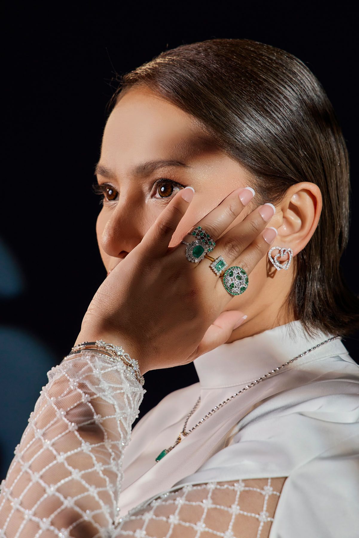 Close-up of woman wearing emerald and diamond rings with sheer lace sleeve against dark background.