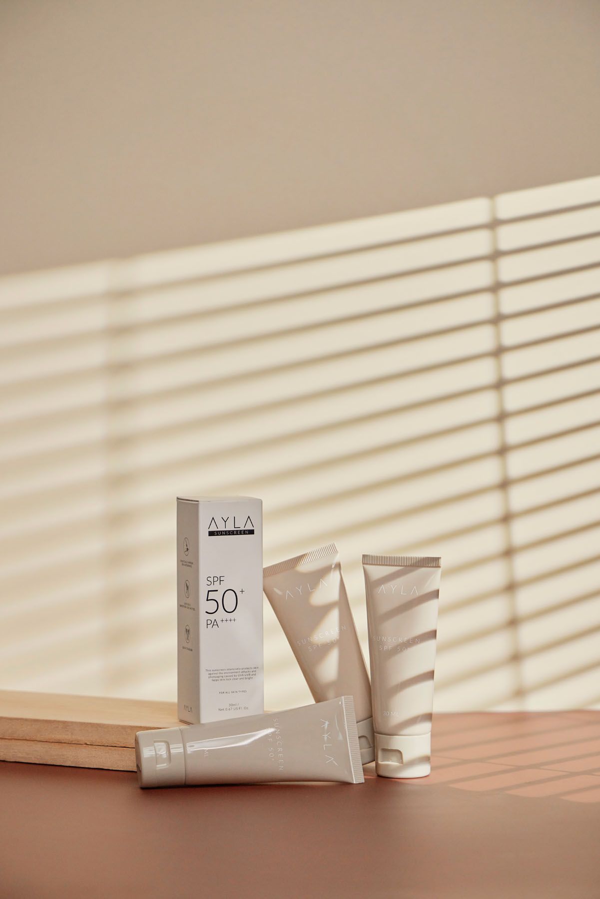 AYLA SPF 50 PA sunscreen tubes and box displayed on wooden surface with soft shadow patterns from window blinds.