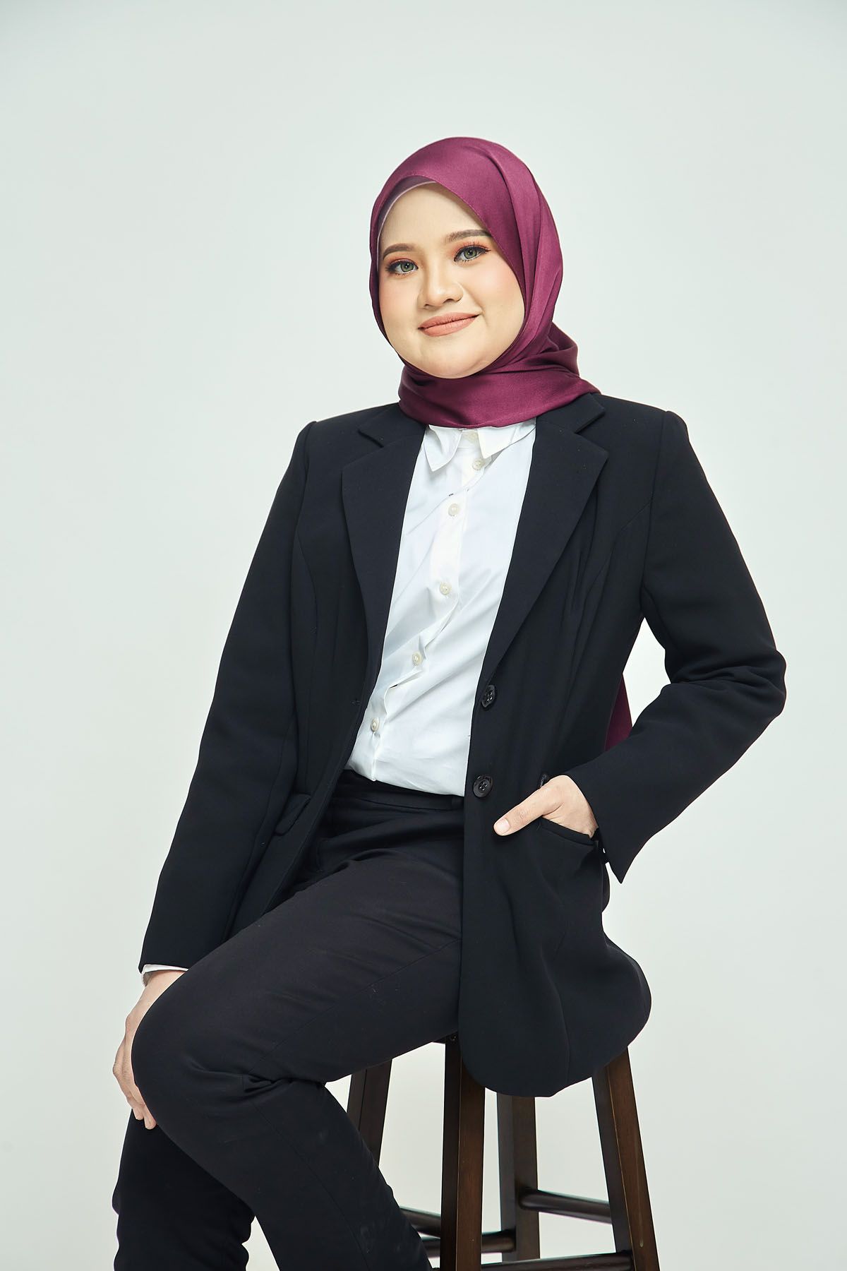Young Muslim woman in black suit and burgundy hijab sitting on wooden stool with stylish pose.