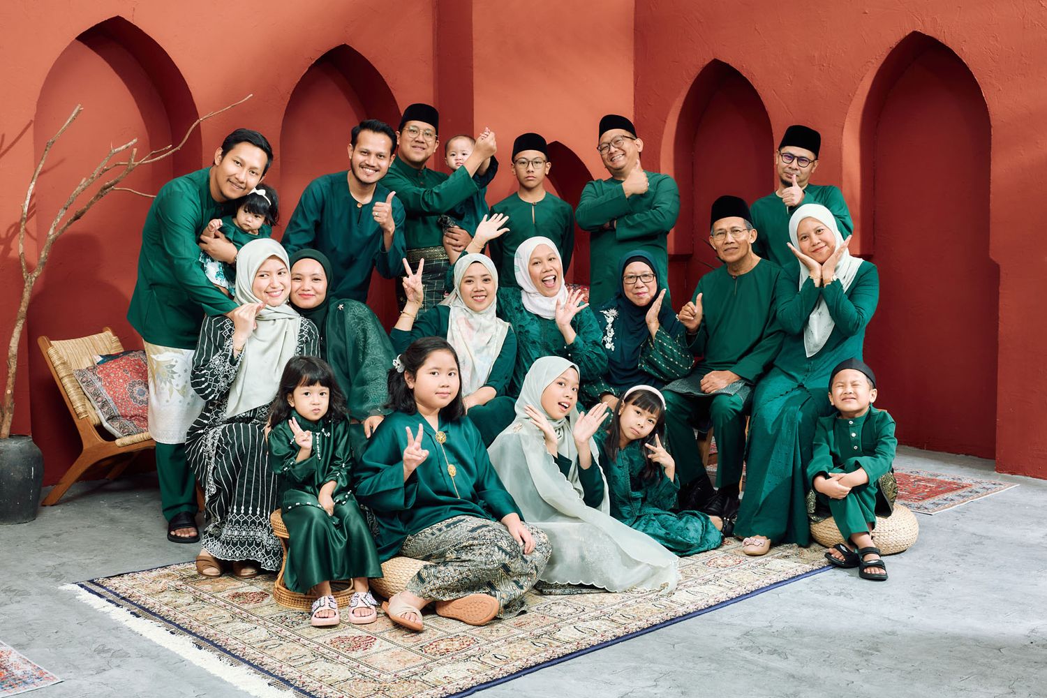 Extended Malaysian family in green traditional Raya outfits smiling together outdoors in a red courtyard.
