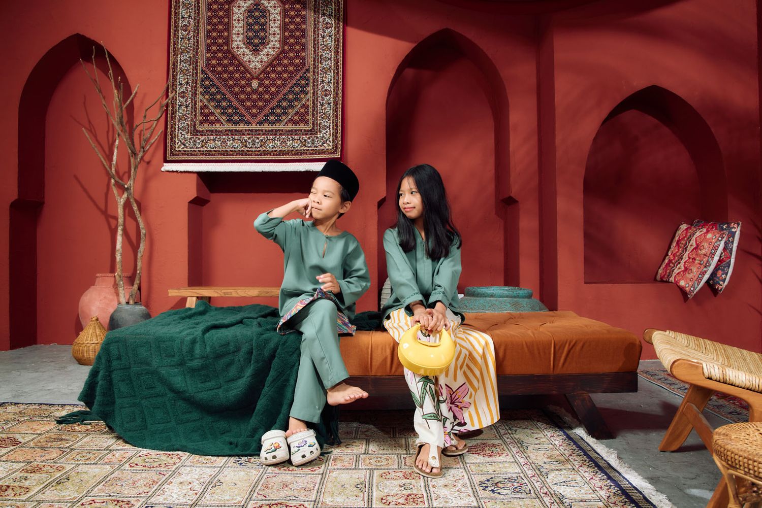Two children in green traditional Malay outfits sitting on a daybed in a decorative red Moroccan-style room.