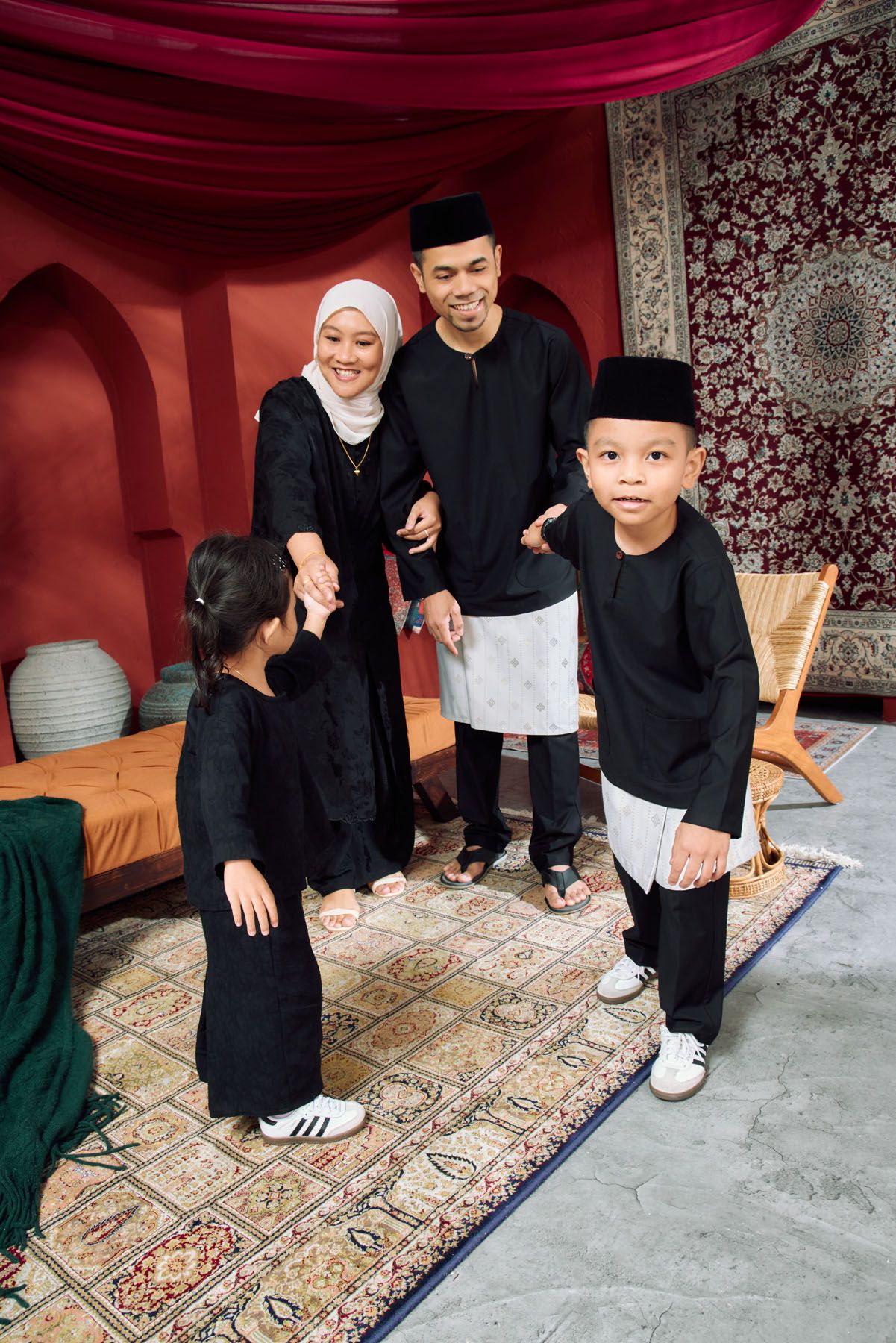 Muslim family in black traditional Malay attire standing together in decorated Eid celebration home.