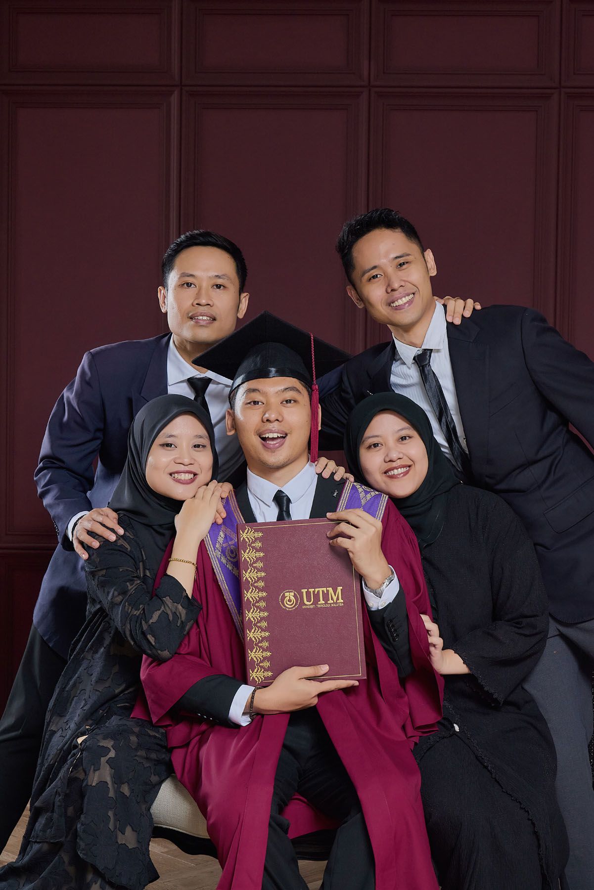 Five family members pose cheerfully with UTM graduate holding diploma in maroon cap and gown.