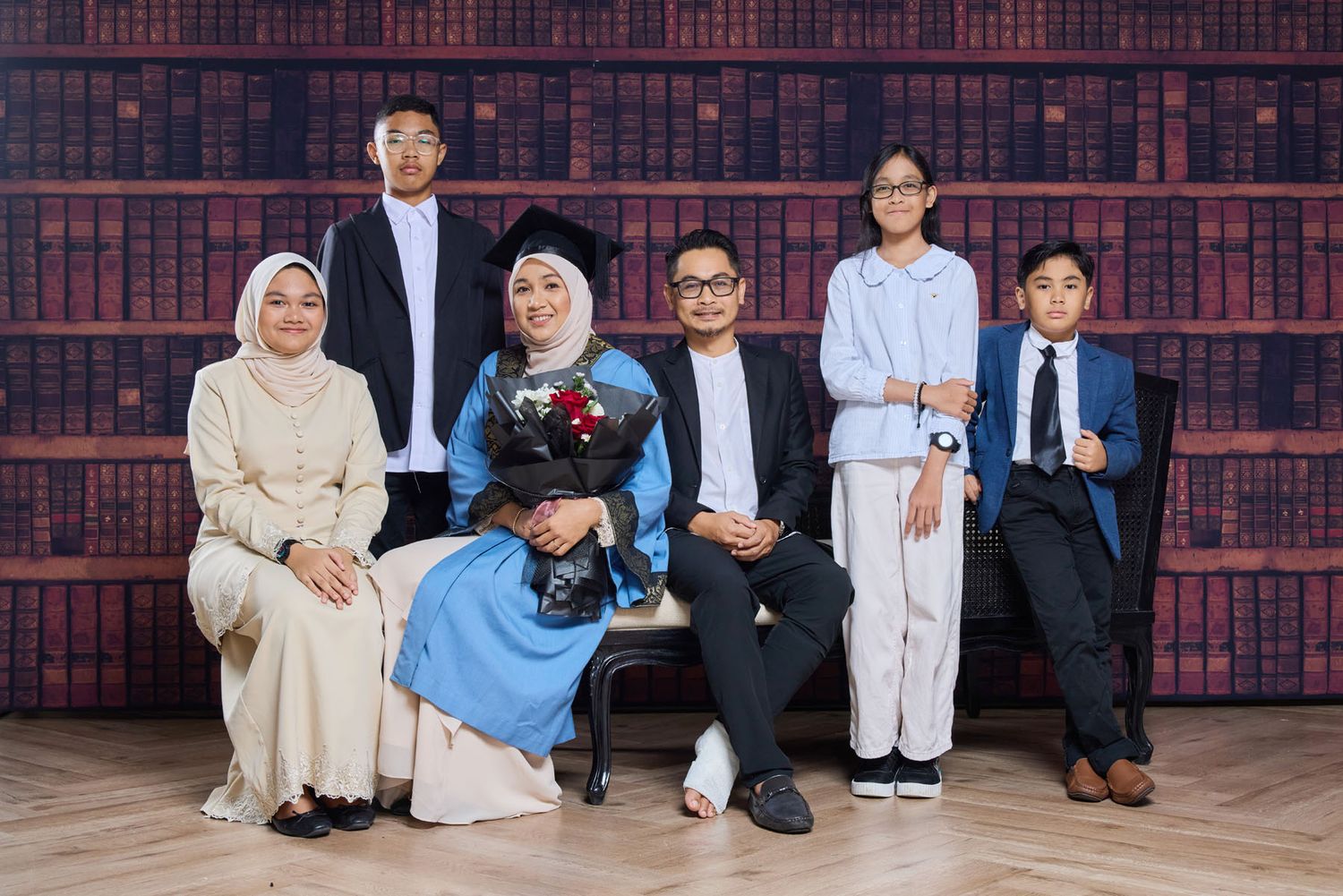 Muslim family of six posing with female graduate in blue gown holding flowers against bookshelf backdrop.