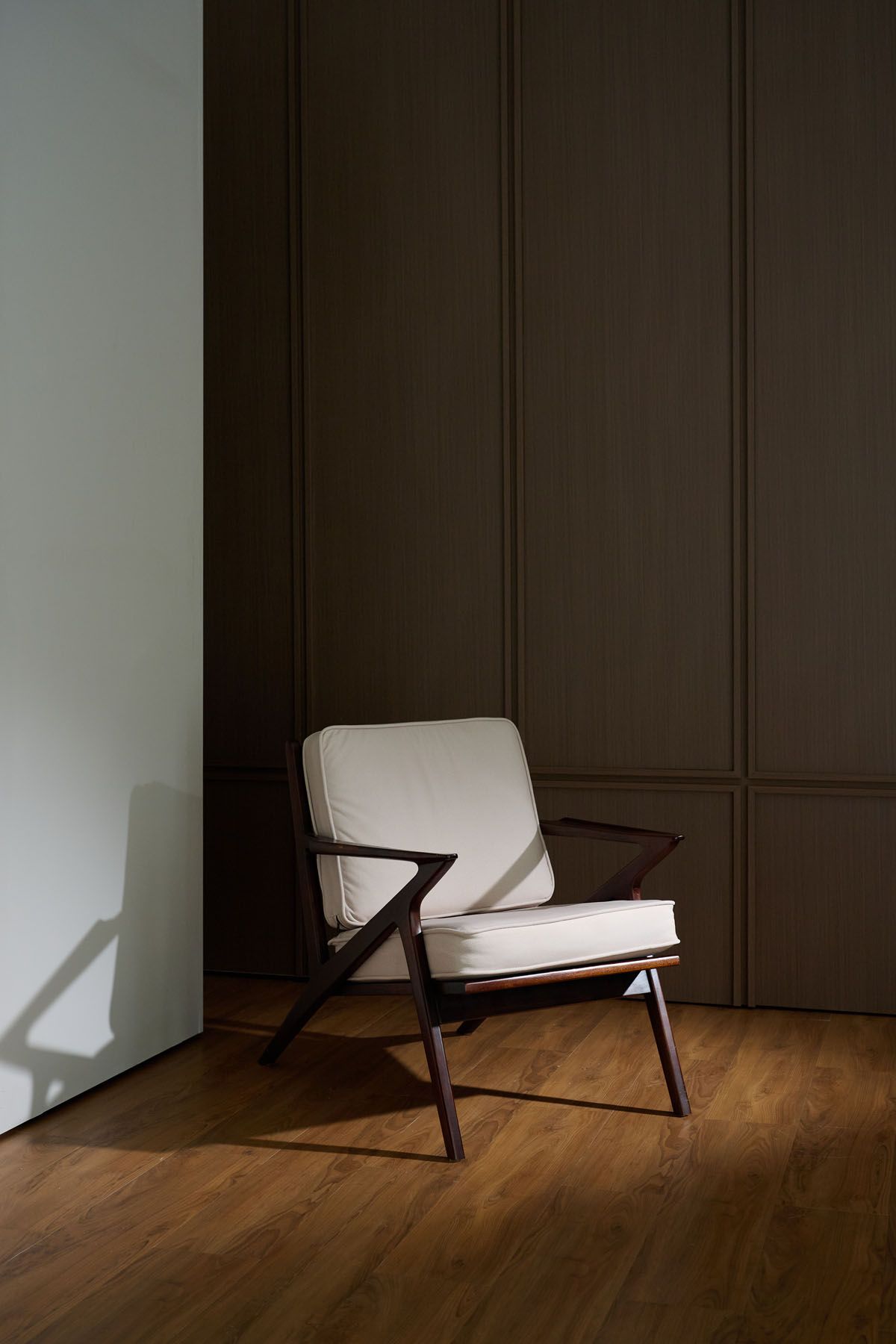 Mid-century modern accent chair with white cushions and dark wood frame on hardwood floor against olive green paneled wall.