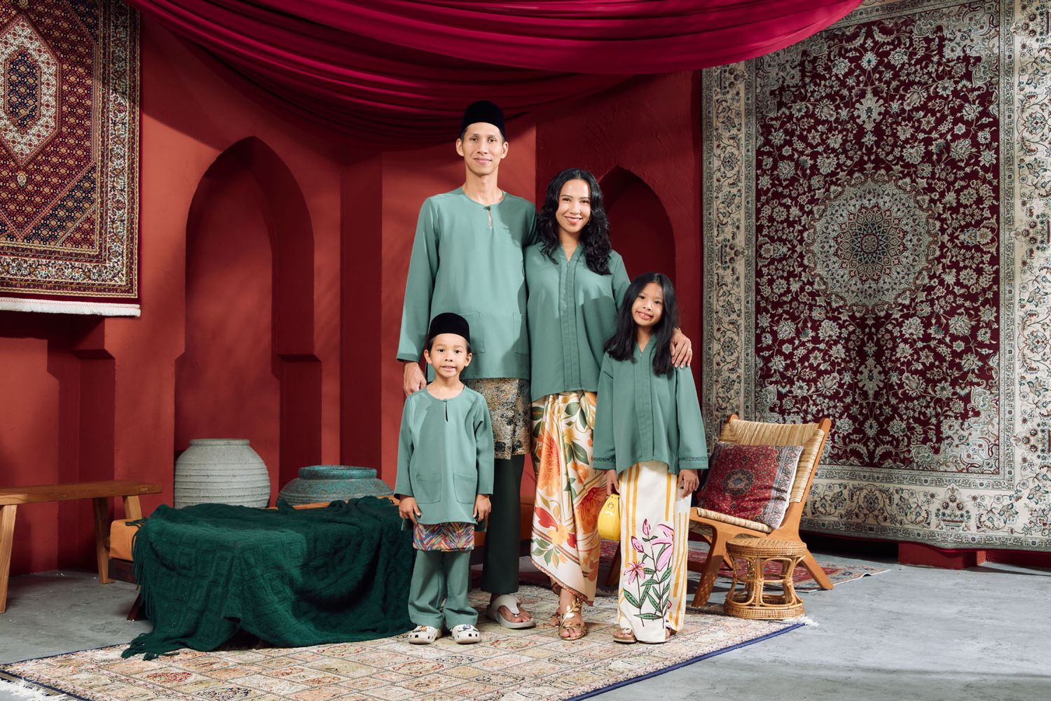 Family of four wearing matching dark green Baju Melayu and Baju Kurung in a Moroccan-inspired red setting.