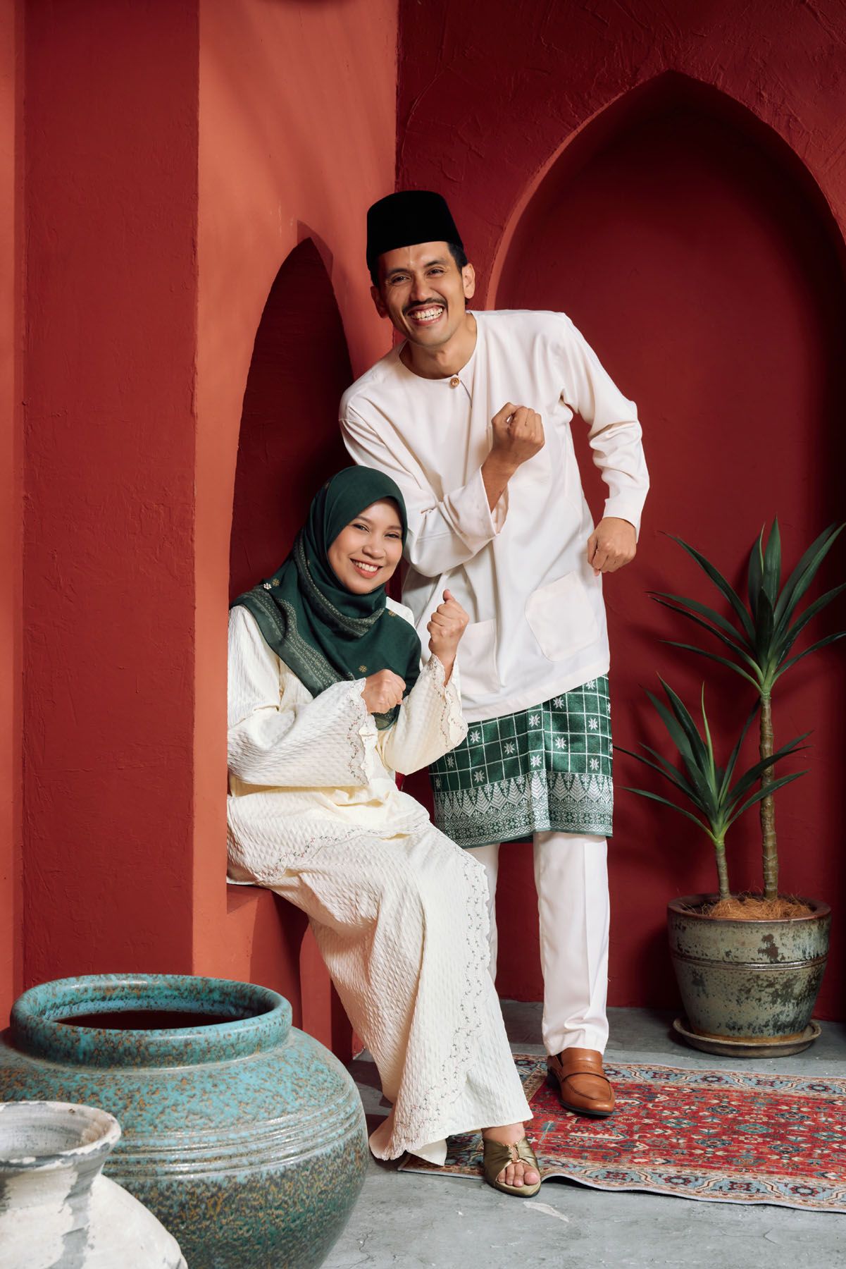 Smiling Muslim couple in traditional Malay attire celebrating Eid in a red archway setting.