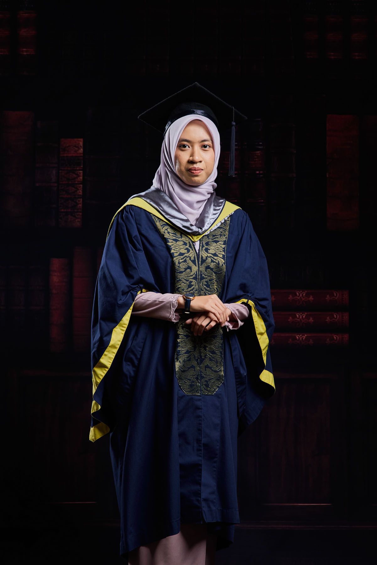 Muslim woman graduate in navy blue robe with yellow trim and hijab posing at ceremony.