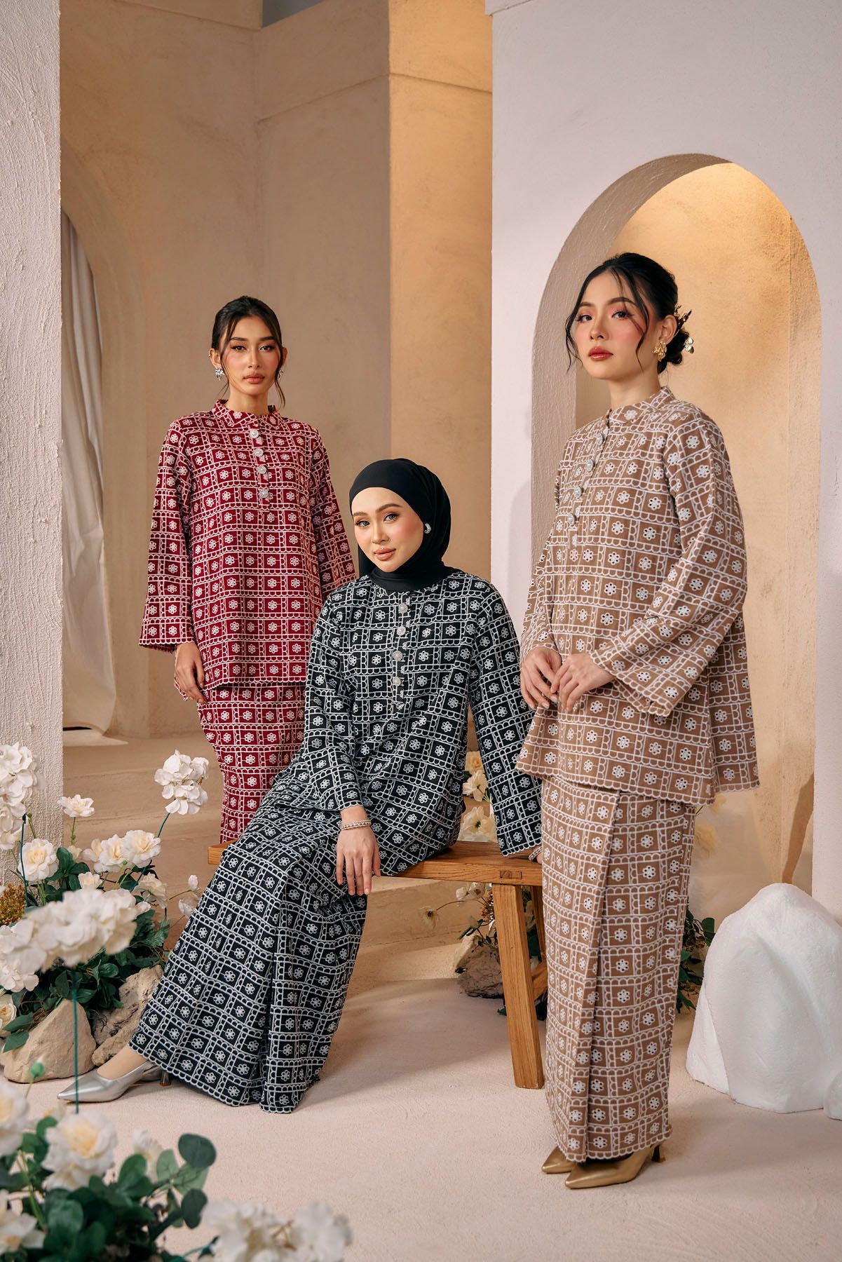 Three women modeling geometric patterned baju kurung in pink, teal, and beige in a styled studio setting.