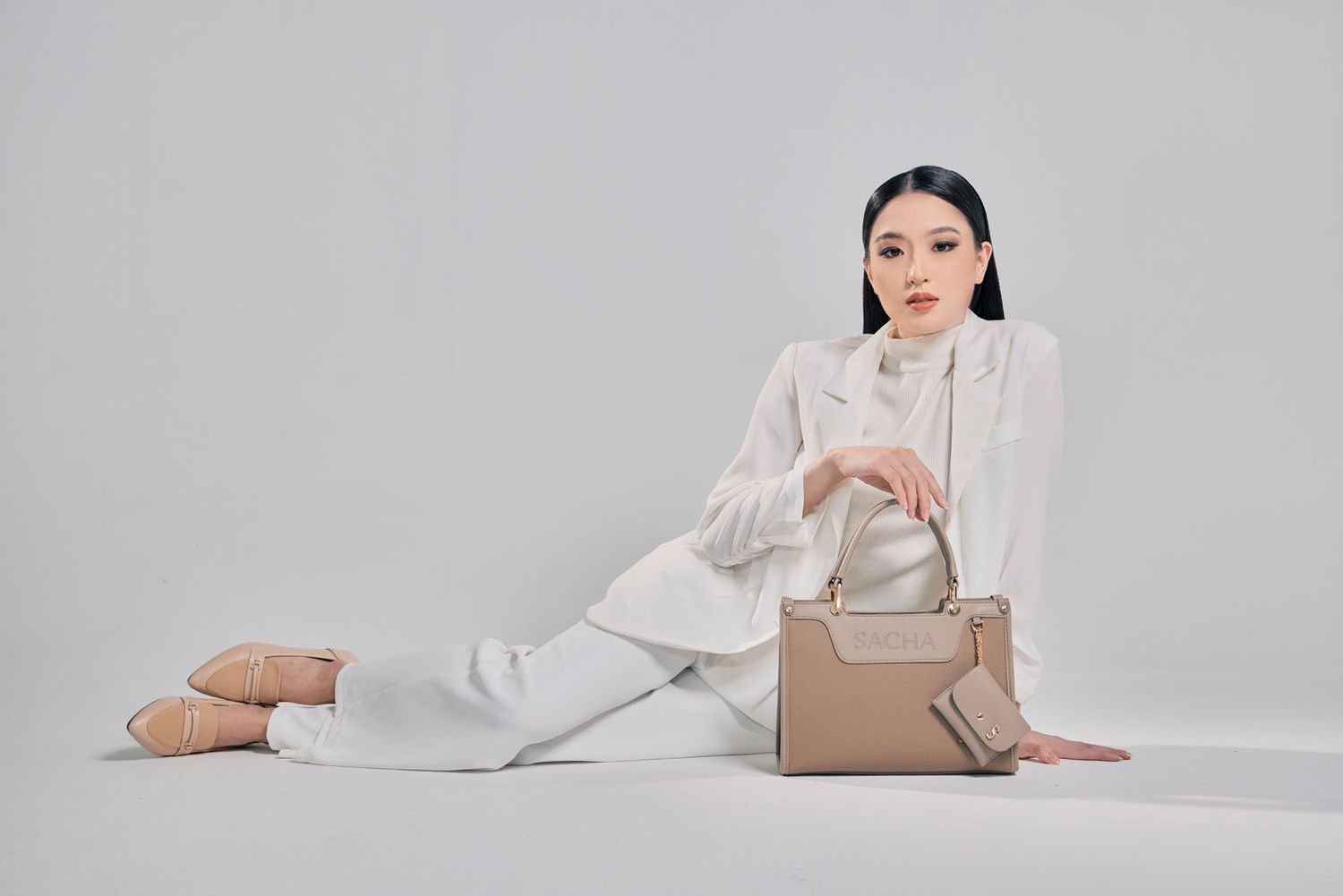 Woman in white blazer sitting on floor with a beige structured handbag and matching wallet.