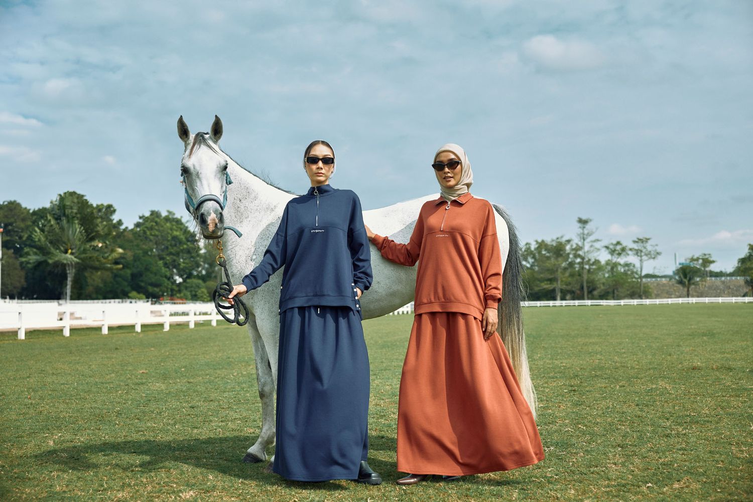 Two women in matching navy and rust skirt sets stand beside a white horse on grass.