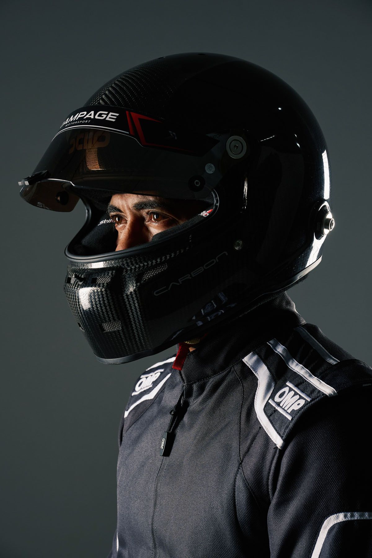 Racing driver in black OMP suit wearing a Rampage carbon fiber helmet, photographed against a dark studio background.