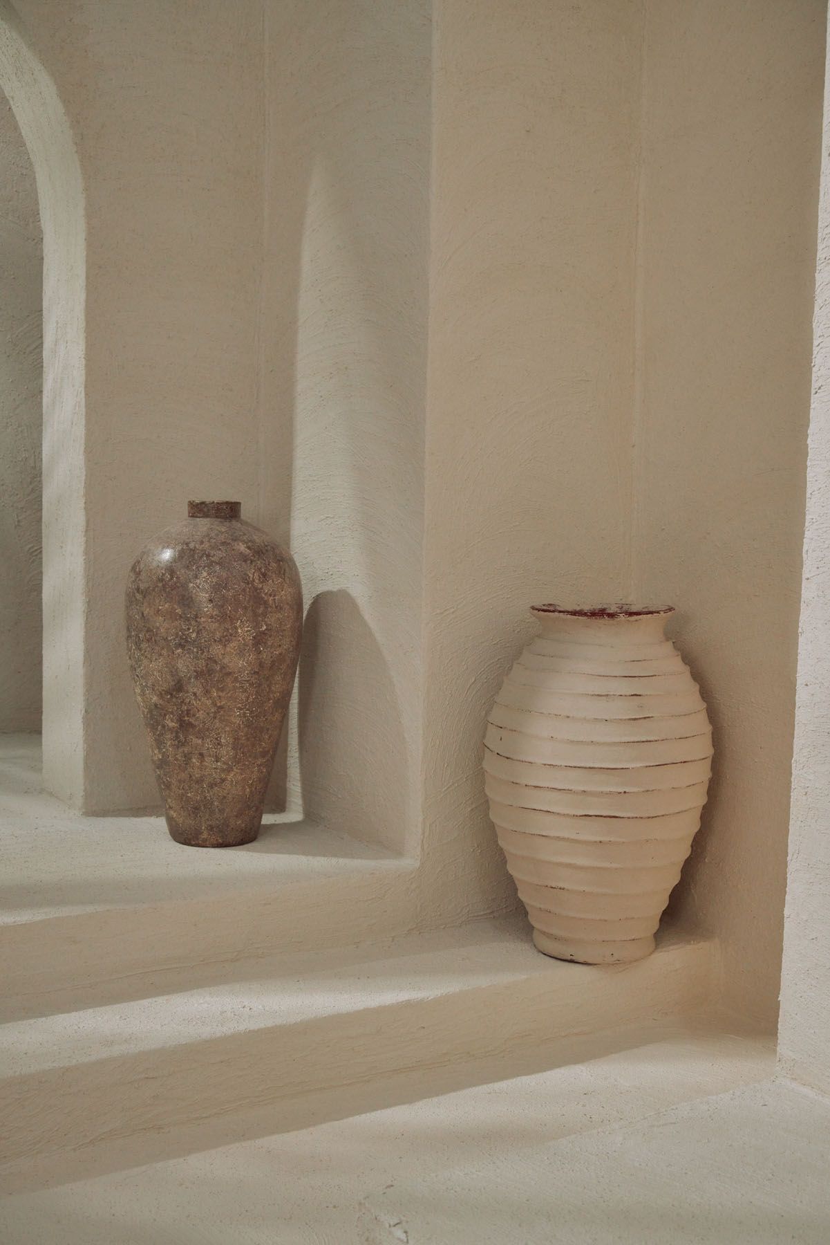 Two rustic clay vases in earthy tones displayed in a minimalist white plaster stepped interior.