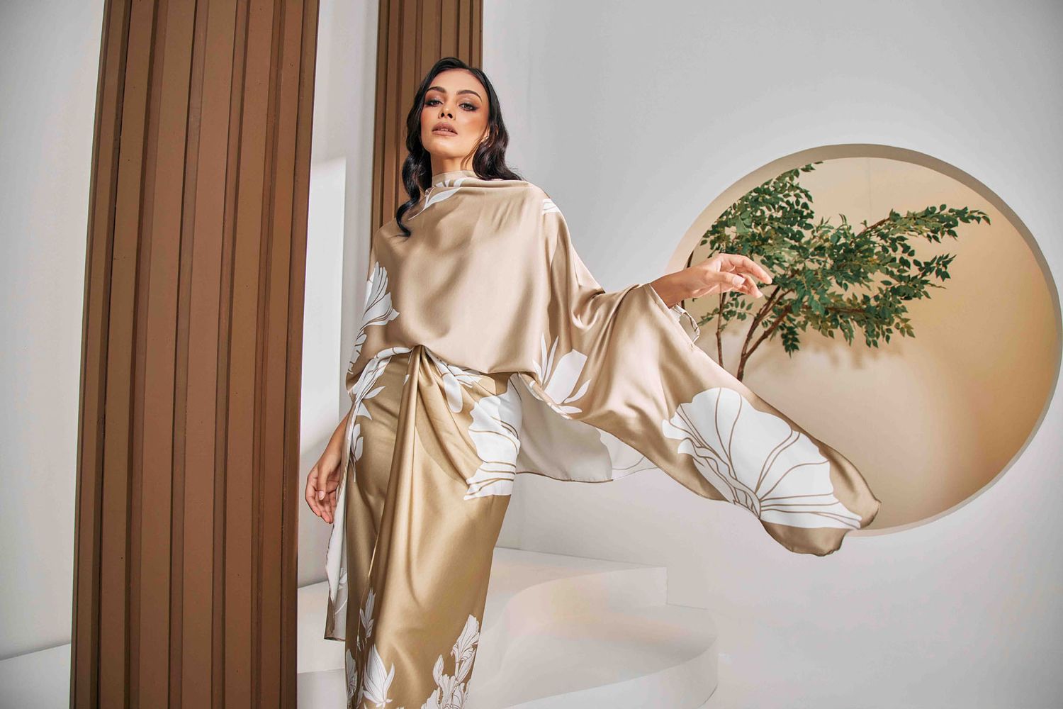 Elegant Muslim woman in gold satin abaya posing near round mirror with green plant decor.
